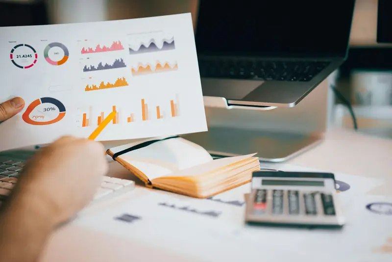 A hand holding a printed page of charts beside a laptop, a notebook and a calculator.