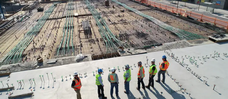 A construction crew in hard hats and safety vests, seen from above, looking over a large site where a foundation is being laid.