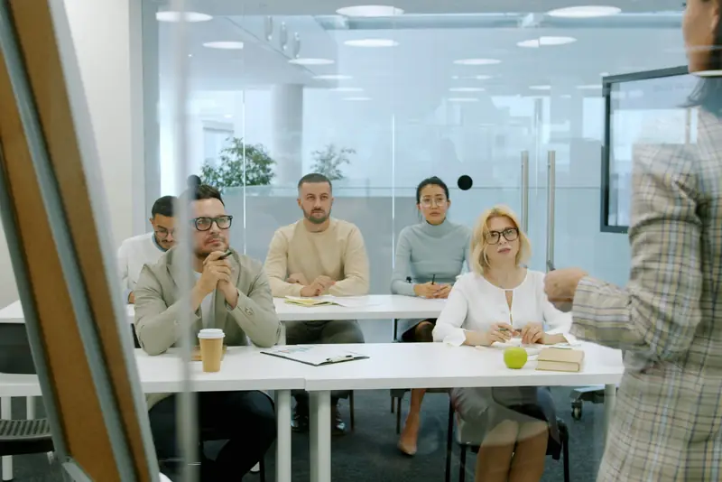 A colleague, seen from behind, presenting to a small group seated at tables in a bright office.