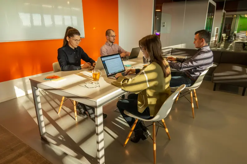 A small team working on laptops around a long table in a warm, sunlit office.